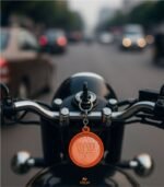 First-person view of bike handle with BJP plastic keychain hanging from ignition key, city traffic blurred in background