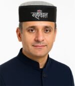 Indian man wearing a black Mahakal Himachali cap with white embroidered text and red tilak symbol.