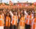 Energetic BJP rally crowd wearing bright orange lotus-print mufflers and waving saffron BJP flags during a large political gathering.