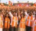 Energetic BJP rally crowd wearing bright orange lotus-print mufflers and waving saffron BJP flags during a large political gathering.