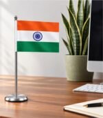 Indian table flag on a silver stand with a round steel base, placed on a wooden desk beside a computer and potted plant.